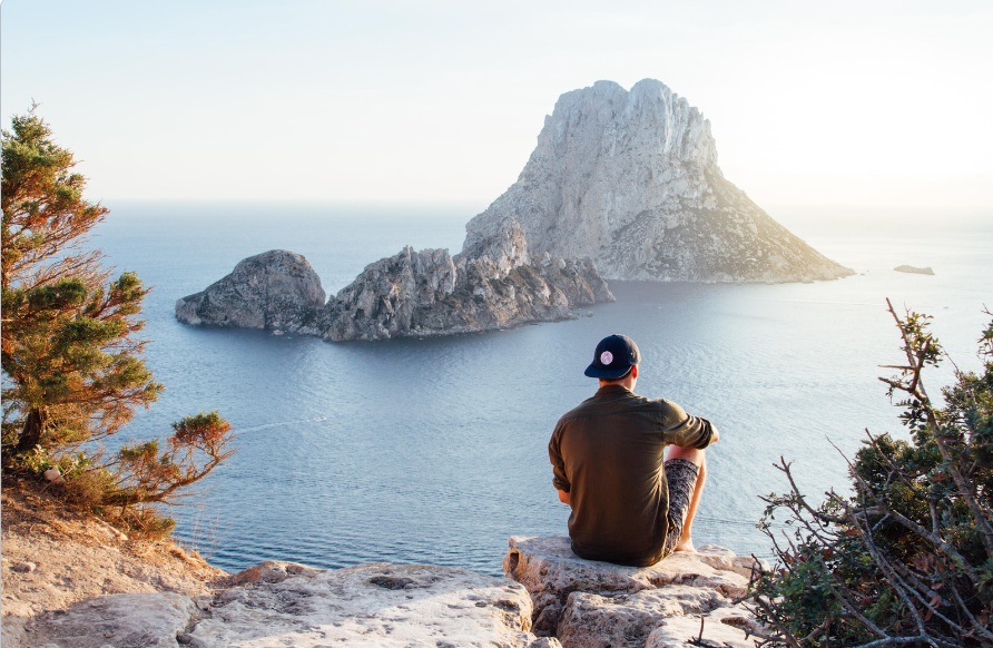 This is an image of a person sitting at a cliff's edge, gazing out over the ocean.